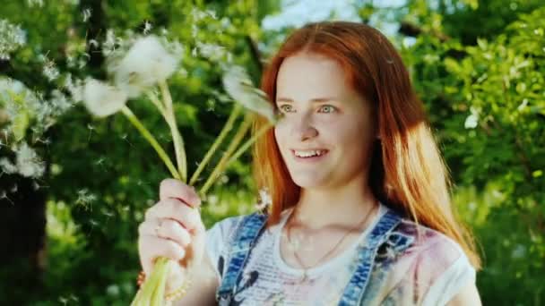 Una chica con el pelo rojo brillante se divierte, juega con flores de diente de león — Vídeos de Stock