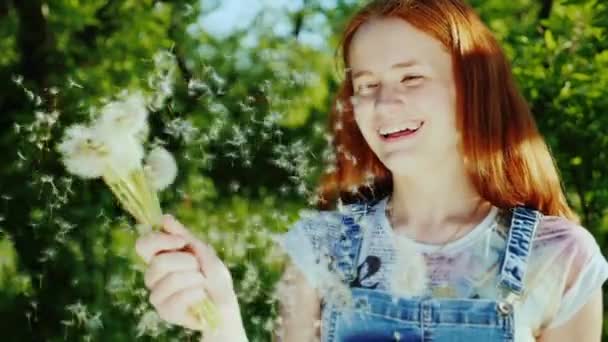 Rolig rödhåriga tonåring flicka som leker med en maskros blomma. Skratt, fröna flyger vackert runt. 180 fps slowmotion video — Stockvideo