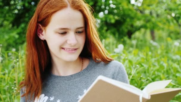 Adolescente pelirroja leyendo un libro en el parque. 4K video en cámara lenta — Vídeo de stock