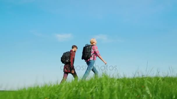 Un uomo e una donna con gli zaini camminano dietro un prato verde sullo sfondo di un bellissimo cielo blu. Viaggi, turismo, una vita sana — Video Stock