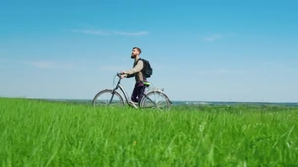 Ecoturismo - um homem dirige uma bicicleta através de um prado verde contra um céu azul brilhante. Uma paisagem idílica, um lugar ambientalmente amigável — Vídeo de Stock