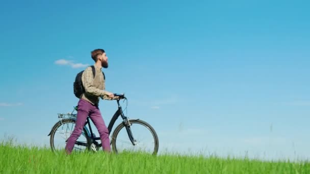 Ένας νεαρός hipster άνδρας οδηγεί ένα ποδήλατο μέσα από ένα πράσινο λιβάδι. Πανέμορφο τοπίο, γραφικό μέρος. — Αρχείο Βίντεο