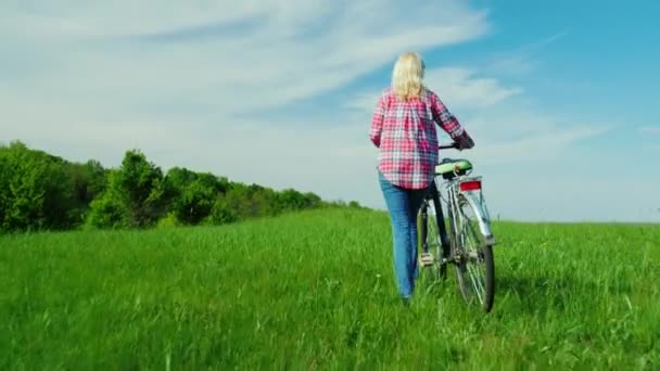 Uma mulher com uma bicicleta caminha em um lugar pitoresco - um prado verde e um céu azul brilhante. Vista traseira. Composoção do espaço de cópia — Vídeo de Stock