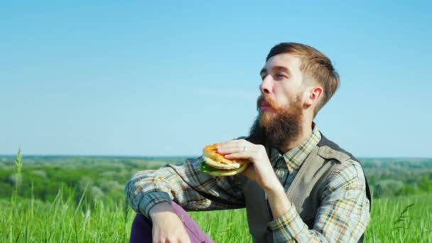 若いあごひげを生やした屋外ハンバーガーを食べるします。スリング晴れた日に景勝地の草原に座っています。 — ストック動画