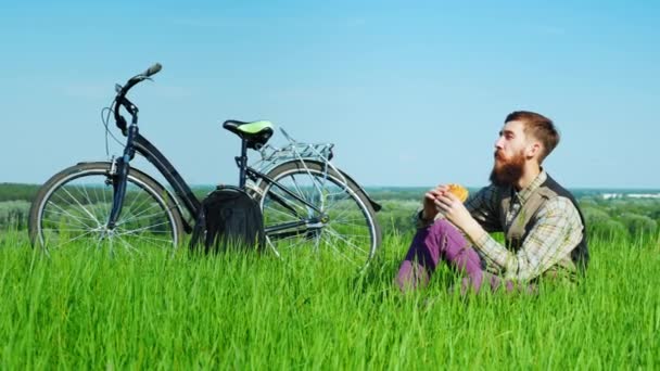 Um lanche na natureza. Jovem comendo um hambúrguer, sentado em um prado verde perto de sua bicicleta — Vídeo de Stock