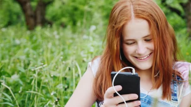 Menina adolescente bonito ruivo desfrutando de um smartphone na natureza. Retrato sentado em um fundo de árvores verdes no parque. Vídeo em câmera lenta 4K — Vídeo de Stock