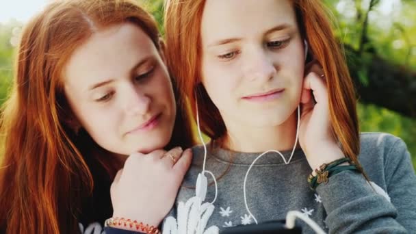 Dos hermanas gemelas de pelo rojo escuchan música en los auriculares. El sol ilumina maravillosamente su cabello. 4K video en cámara lenta — Vídeo de stock