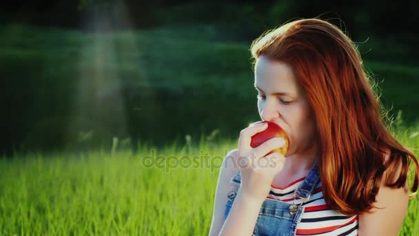 Adolescente dai capelli rossi che mangia una mela rossa succosa a un picnic. Un posto pittoresco - un prato verde, un bagliore bellissimo dal sole — Video Stock