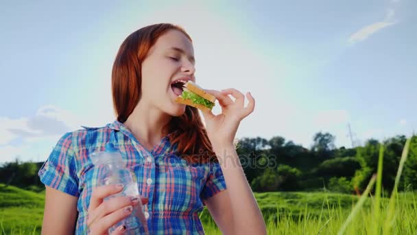 Vörös hajú tinédzser enni egy szendvicset, egy piknik. Tart egy üveg víz a kezében — Stock videók