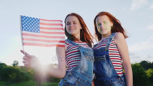 Dos chicas adolescentes pelirrojos lindos con una bandera americana. Sonríe, mira la cámara. 4K video en cámara lenta — Vídeo de stock