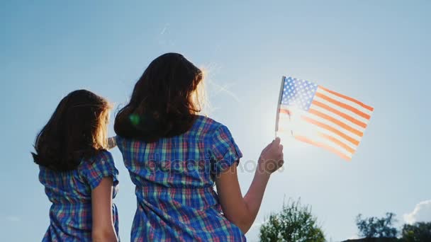 Två unga kvinnor med den amerikanska flaggan. I strålar solen mot himlen. Bakifrån. Självständighetsdagen — Stockvideo