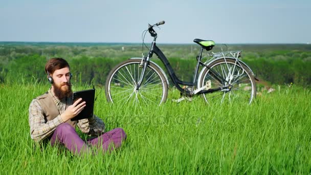 A young attractive man in headphones uses a tablet. Sits on a green meadow or lawn near his bicycle. Always online — Stock Video