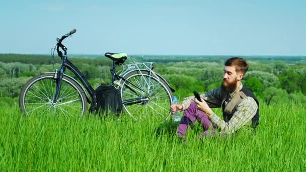En ung biker vilar på en grön äng. Använder en mobiltelefon och dricker vatten ur en flaska — Stockvideo