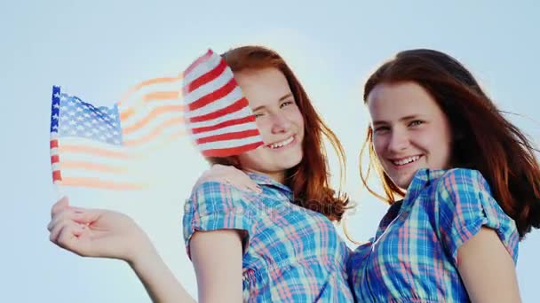 Retrato de gemelas con bandera americana. Mirando a la cámara contra el cielo azul, sonriendo. Día de la independencia — Vídeos de Stock