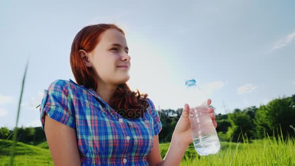 Un adolescente beve acqua pulita da una bottiglia su un picnic in un posto pittoresco. Bellissimo prato verde e cielo blu — Video Stock