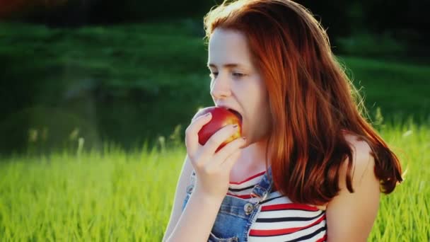 一名少女的画像。在一片绿色的草地上吃一个大的红苹果 — 图库视频影像