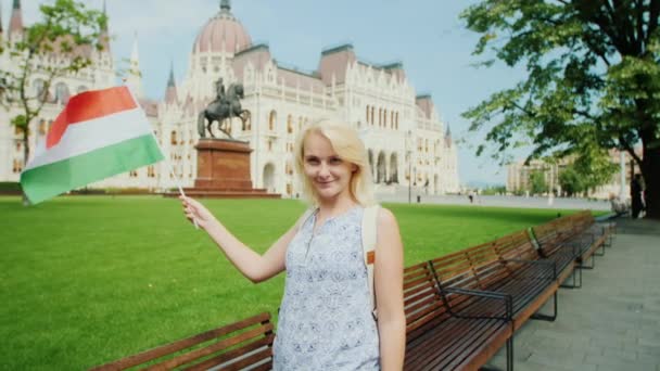 Mujer joven turista posando con la bandera húngara en el fondo del parlamento en Budapest. Turismo en Europa — Vídeos de Stock