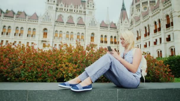 La mujer se sienta en el fondo del parlamento húngaro, utiliza un teléfono inteligente. Turismo en Europa, magnífico Budapest — Vídeo de stock