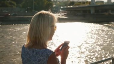 Genç bir kadın, bir akıllı telefon kullanır bir nehir üzerinde bir gemi yola çıkacak. Gün batımından önce arka planda bir parlama fonunda. Arkadan Görünüm