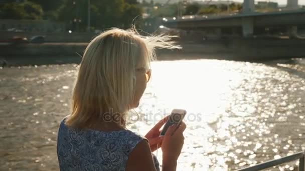 Une jeune femme utilise un smartphone, navigue sur une rivière sur un bateau. Dans le contexte d'un éblouissement en arrière-plan avant le coucher du soleil. Vue arrière — Video