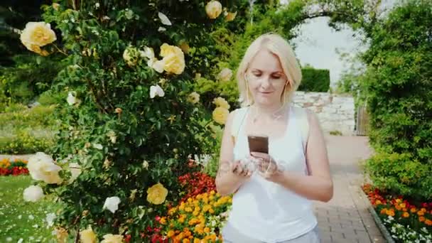 一个年轻的女人正走在花园里的玫瑰。使用移动电话。格拉茨，奥地利 — 图库视频影像