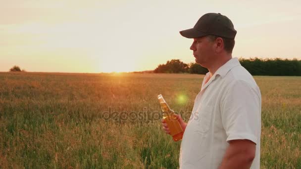 Отдохнуть после тяжелой работы - фермер пьет пиво из бутылки. Стоя на пшеничном поле на закате — стоковое видео