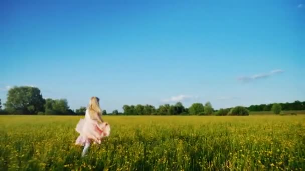 Bekymmerslös tjej i en rosa klänning löper tvärs över fältet med gula blommor. Steadicam slowmotion skott — Stockvideo