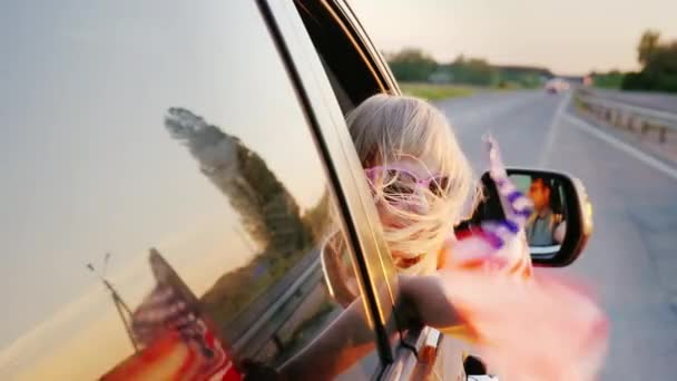 Une petite fille en lunettes roses regarde par la fenêtre de la voiture. Dans sa main tient le drapeau américain — Video