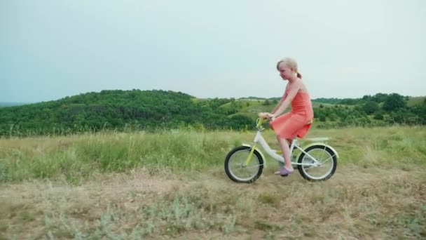 Uma menina em um vestido rosa monta uma bicicleta no campo. Feliz infância despreocupada. Steadicam tiro — Vídeo de Stock