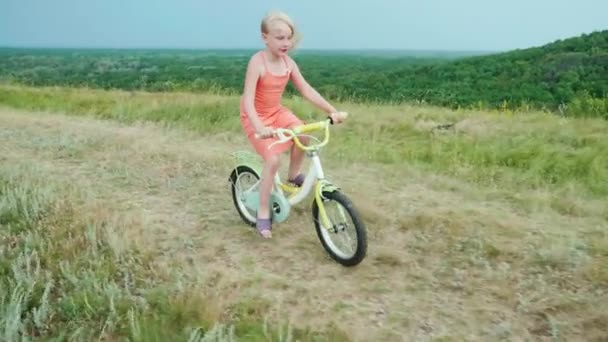 Een cool meisje in een roze jurk rijdt een fiets op het platteland. Zorgeloze kindertijd in het dorp. Slow motion 4k video — Stockvideo