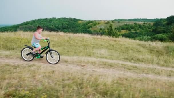 Ένα μικρό αγόρι έρχεται κάτω από ένα λόφο σε ένα ποδήλατο, χαμογελώντας. Ξένοιαστα παιδικά χρόνια και στις αγροτικές περιοχές — Αρχείο Βίντεο
