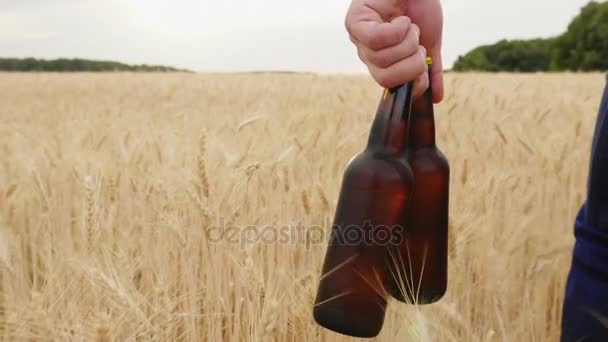 Un hombre lleva dos botellas de cerveza fría sobre un campo de cebada. El concepto es saciar tu sed. Vídeo en cámara lenta — Vídeos de Stock