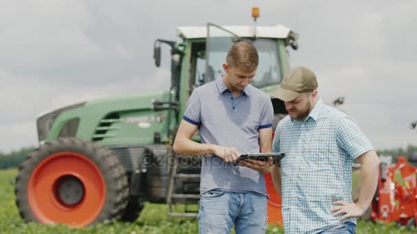 Két fiatal férfi, a mezőgazdasági termelő kommunikál, használja a tabletta. Áll szemben a traktor háttér területén — Stock videók