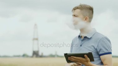 Genç bir adam elektronik sigara içiyor, bir tablet kullanır. Bir petrol sondaj platformu fonunda