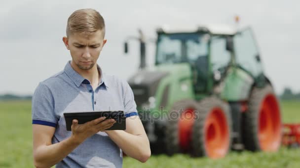 Un joven agricultor está trabajando en el campo. Utiliza una tableta, en el fondo hay un tractor grande — Vídeo de stock