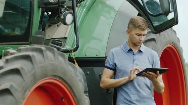 Un joven conductor de tractor utiliza una tableta. Un granjero está cerca de una máquina agrícola — Vídeo de stock