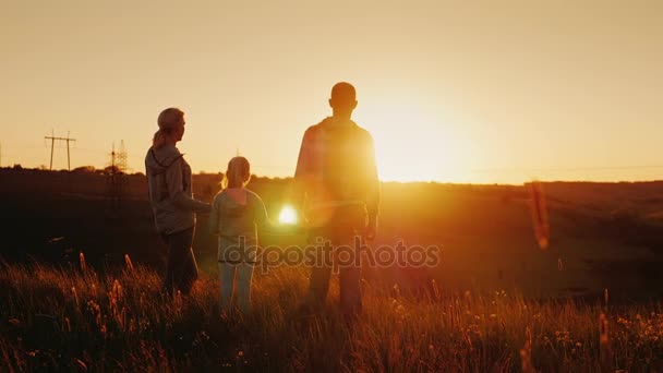 Uma família jovem mãe, pai e filha juntos olham para o pôr do sol. Felizes juntos, silhuetas ao pôr-do-sol. Visão traseira — Vídeo de Stock