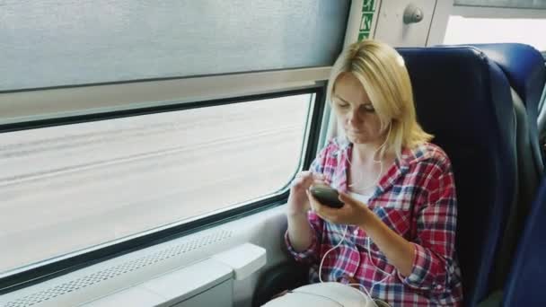Una mujer está viajando en el segundo piso de un tren, sentada junto a la ventana. Utiliza un teléfono inteligente en la carretera — Vídeo de stock