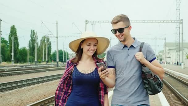 Glada unga par turister med en smartphone går till tågstationen. Koncept: Beställ online biljetter — Stockvideo