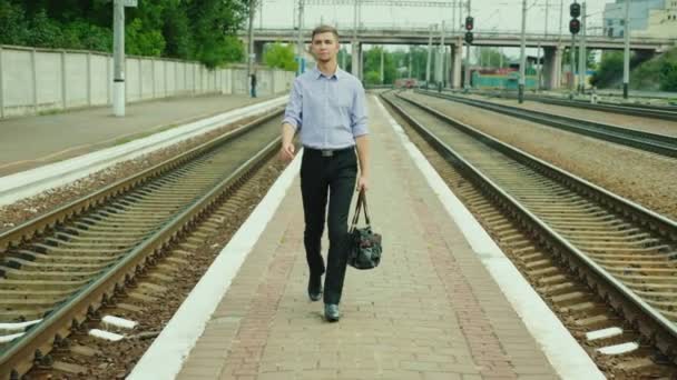 若いビジネスマンは、トラックに沿って駅を歩きます。彼は彼の手、笑顔で袋を運ぶ。成功するビジネス旅行 — ストック動画