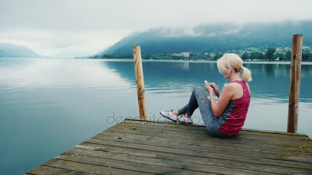 Siempre conectado. Una mujer utiliza un teléfono inteligente, se sienta en un lugar pintoresco cerca de un lago de montaña en los Alpes — Vídeo de stock