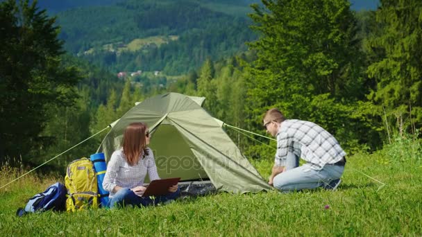 Los amigos están armando una tienda. Una mujer está usando un portátil, un hombre está revisando cuerdas. Camping en el camping — Vídeos de Stock