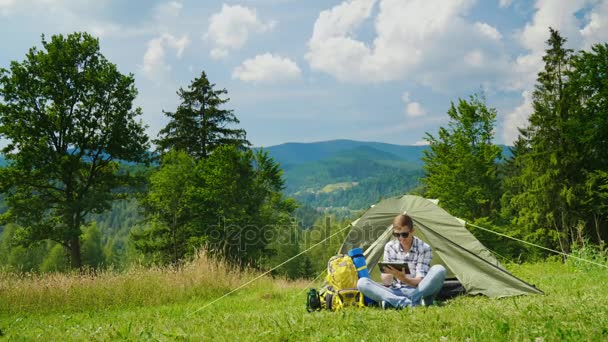 Un joven turista masculino usa una tableta en el campamento cerca de la tienda. En un lugar pintoresco en el fondo de las montañas. Composición del espacio de copia — Vídeos de Stock