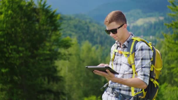 バックパックで若い男性の観光客は、タブレットを使用しています。それは山の近くの美しい場所に立っています。 — ストック動画