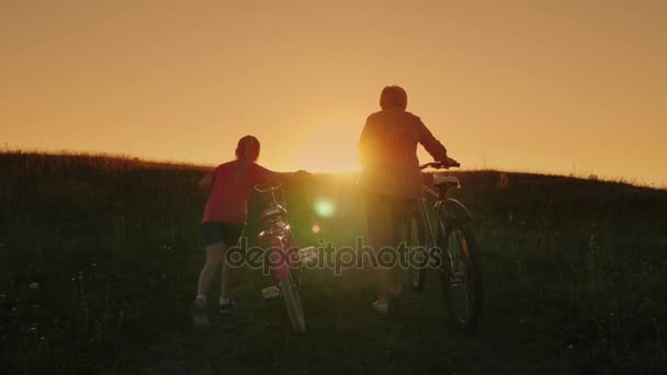 Aktive ältere Menschen. Eine ältere Frau besteigt mit ihrer Enkelin einen Berg und führt Fahrräder. Rückansicht bei Sonnenuntergang — Stockvideo