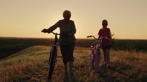 Uma mulher idosa está andando com sua neta no campo. Eles estão andando ao lado, dirigindo bicicletas. Conceito - pessoas idosas ativas — Vídeo de Stock