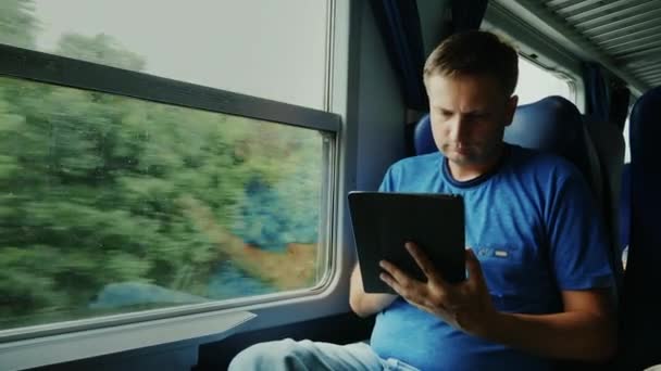 Un uomo sta viaggiando su un treno. Si siede vicino alla finestra, usa un tablet digitale. Dispositivo sulla strada — Video Stock