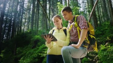 Turist sırt çantaları ile mutlu çift ormanda kendilerini yönlendirmek, bir tablet kullanın. Güzel sabah güneş ışınlarının onları aydınlatmak.