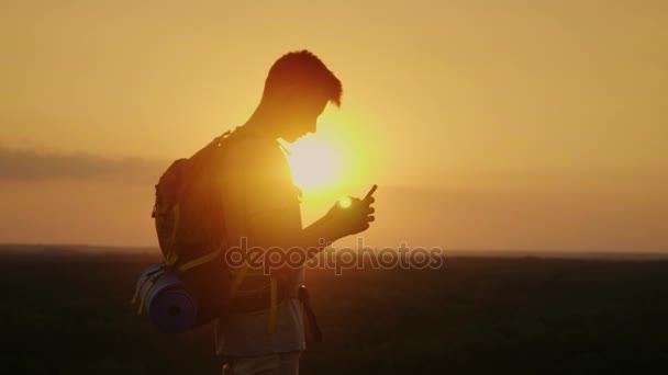 젊은 관광객 남자는 배낭에 산 위에 휴대 전화를 사용합니다. 주황색 하늘 석양의 광선. 항상 온라인 — 비디오