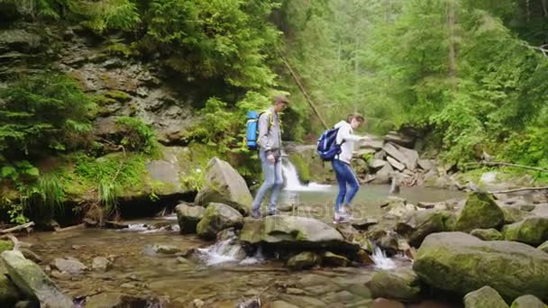 Una joven pareja de turistas con mochilas cruza un río de montaña en el bosque — Vídeos de Stock